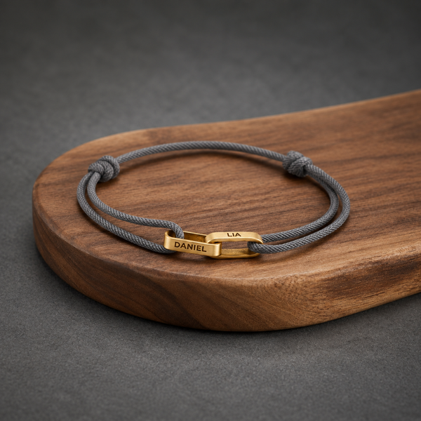 Personalised couple bracelet on a grey cord, two gold bars engraved with a name each, on a wooden board